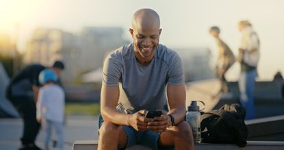 An athletic man at a park smiles and looks at his phone.