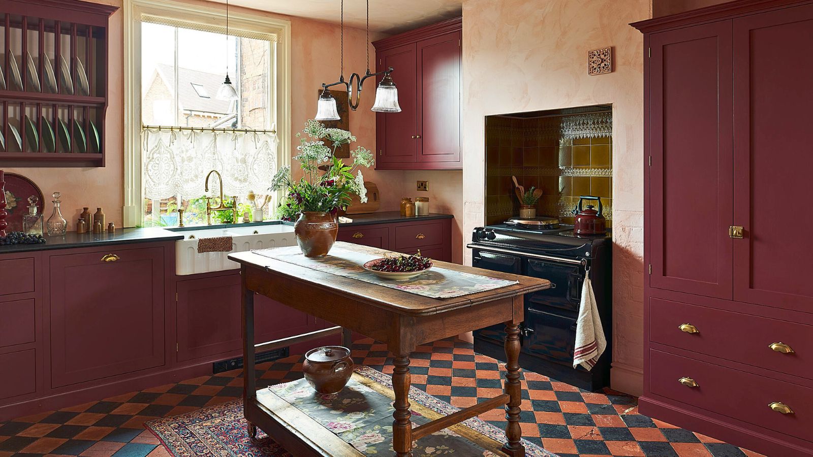 Kitchen with wooden mid-century island, Range Cooker and burgundy built-ins, large window with lace window treatment, open wall-mounted plate rack and orange and navy tiled floor