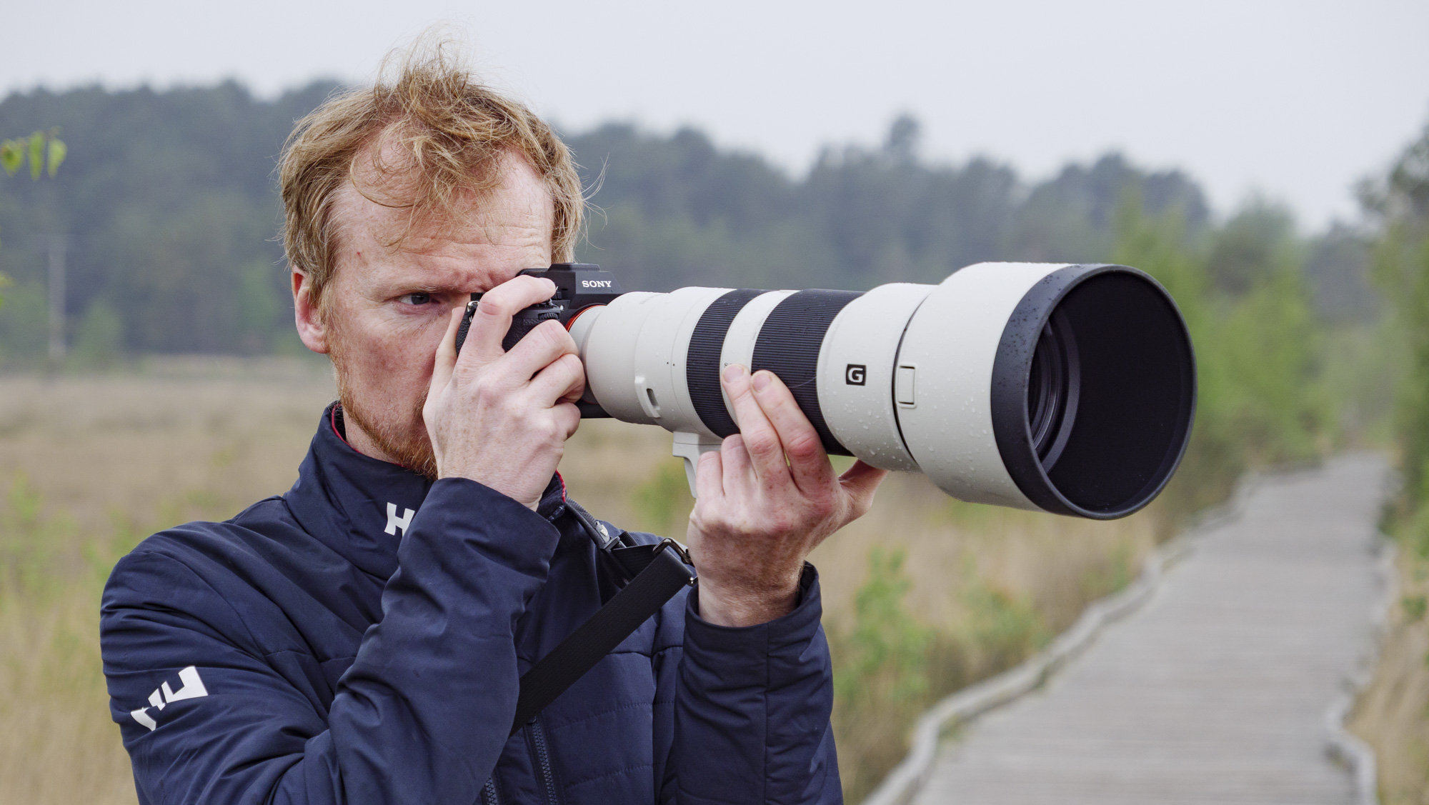 Man holding the Sony A7R VI up to his eye with the FE 400-800mm F6.3-8 G OSS supertelephoto zoom lens attached, he's standing on a boardwalk in a nature reserve