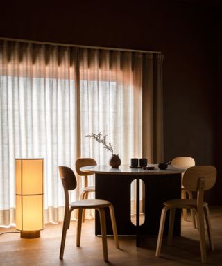 A dimly lit dining room with a large glowy standing lamp and floor-to-ceiling windows covered by curtains