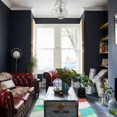 A dark navy-painted living room with high ceilings and a brown leather Chesterfield sofa and matching armchair