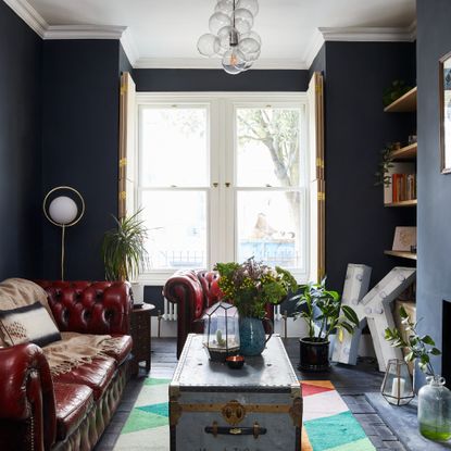 A dark navy-painted living room with high ceilings and a brown leather Chesterfield sofa and matching armchair
