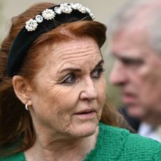 Sarah Ferguson wearing a green coat and black crystal headband looking to the right while Prince Andrew is blurry behind her