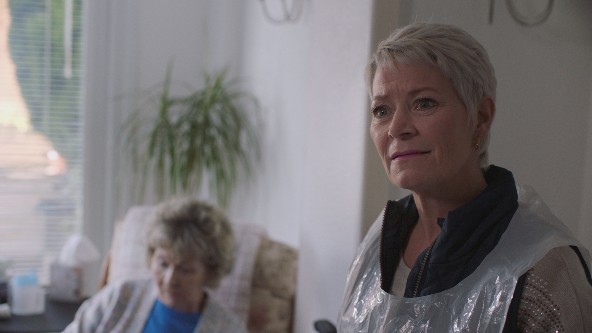 Sharon Percy as Tina Piper, wearing a plastic disposable apron over her clothes and smiling, as an older woman sits on an armchair in the background.