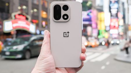 OnePlus 15 in Sand Storm color held in hand with cameras and back showing in the middle of a Times Square New York City intersection across from the Hard Rock Hotel. The background is blurred with cars and people moving past