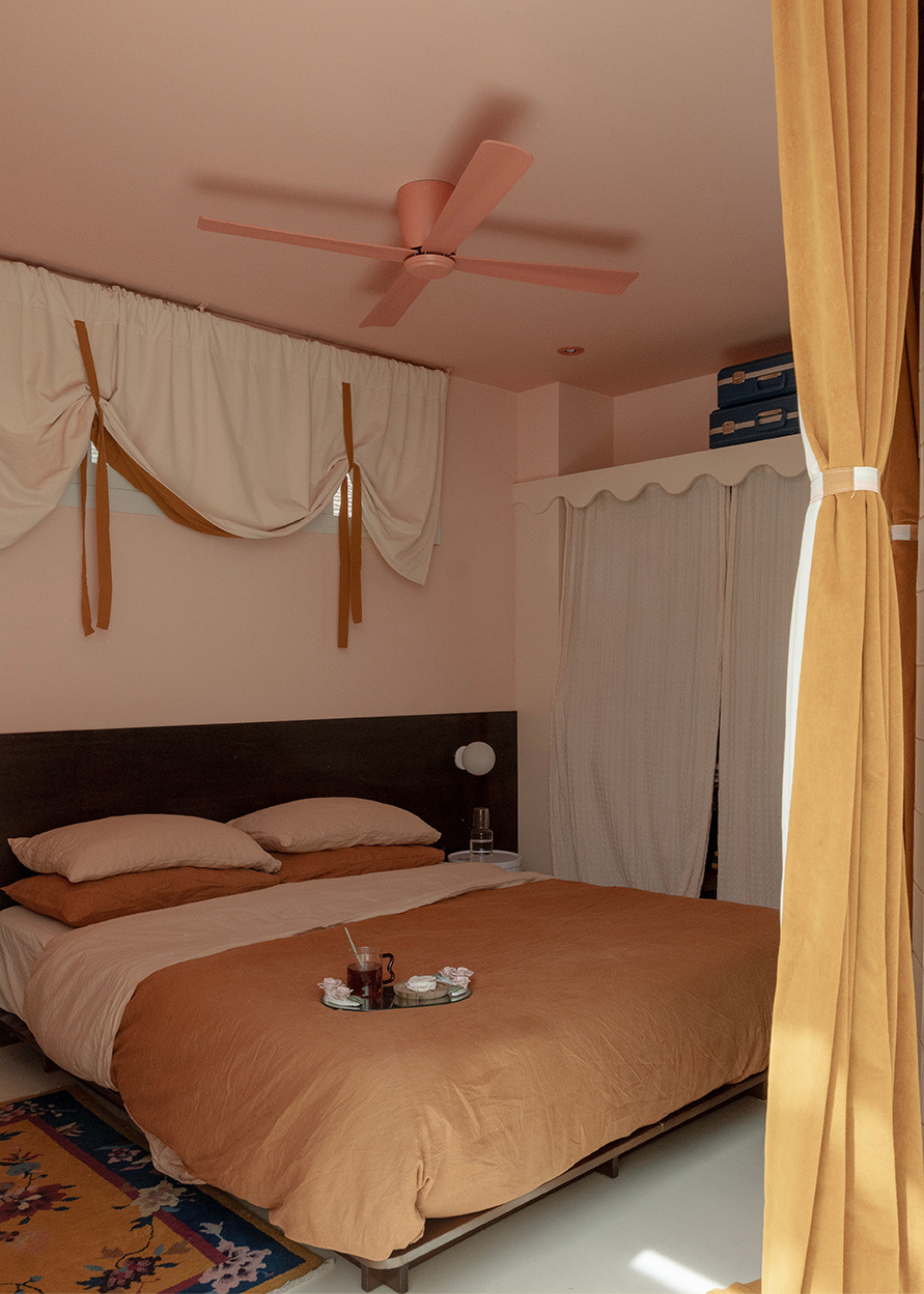 A pale pink bedroom with a pink ceiling fan, a low brown bed with a tray of drinks, by a patterned rug