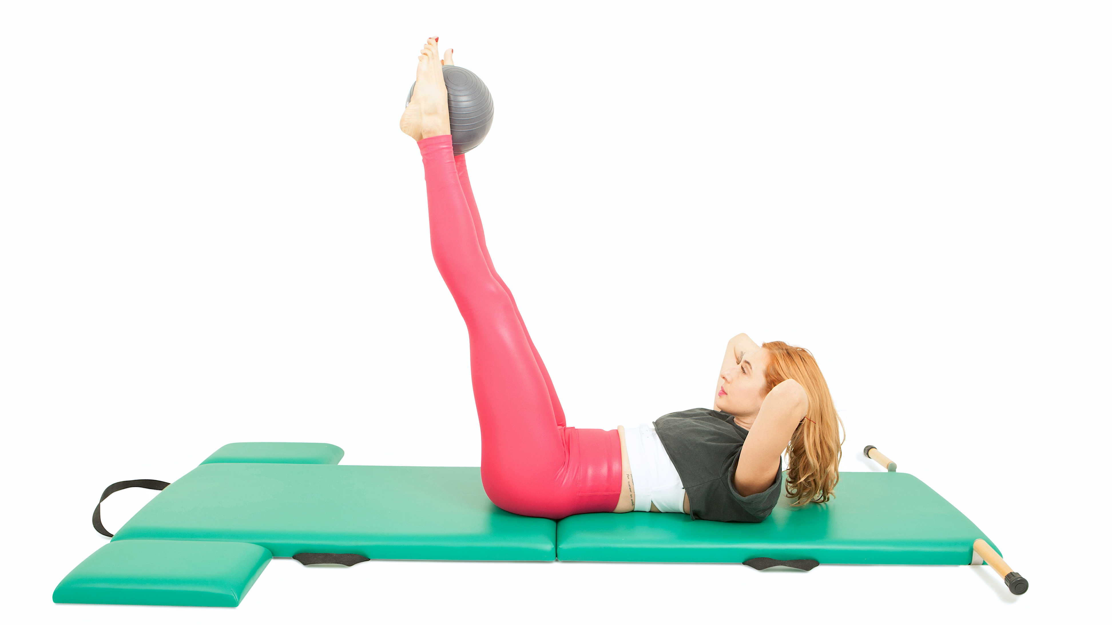Lesley Logan lies on a padded mat with her feet and legs elevated, back against the mat, and hands behind her elevated head. She squeezes a Pilates ball between her feet.