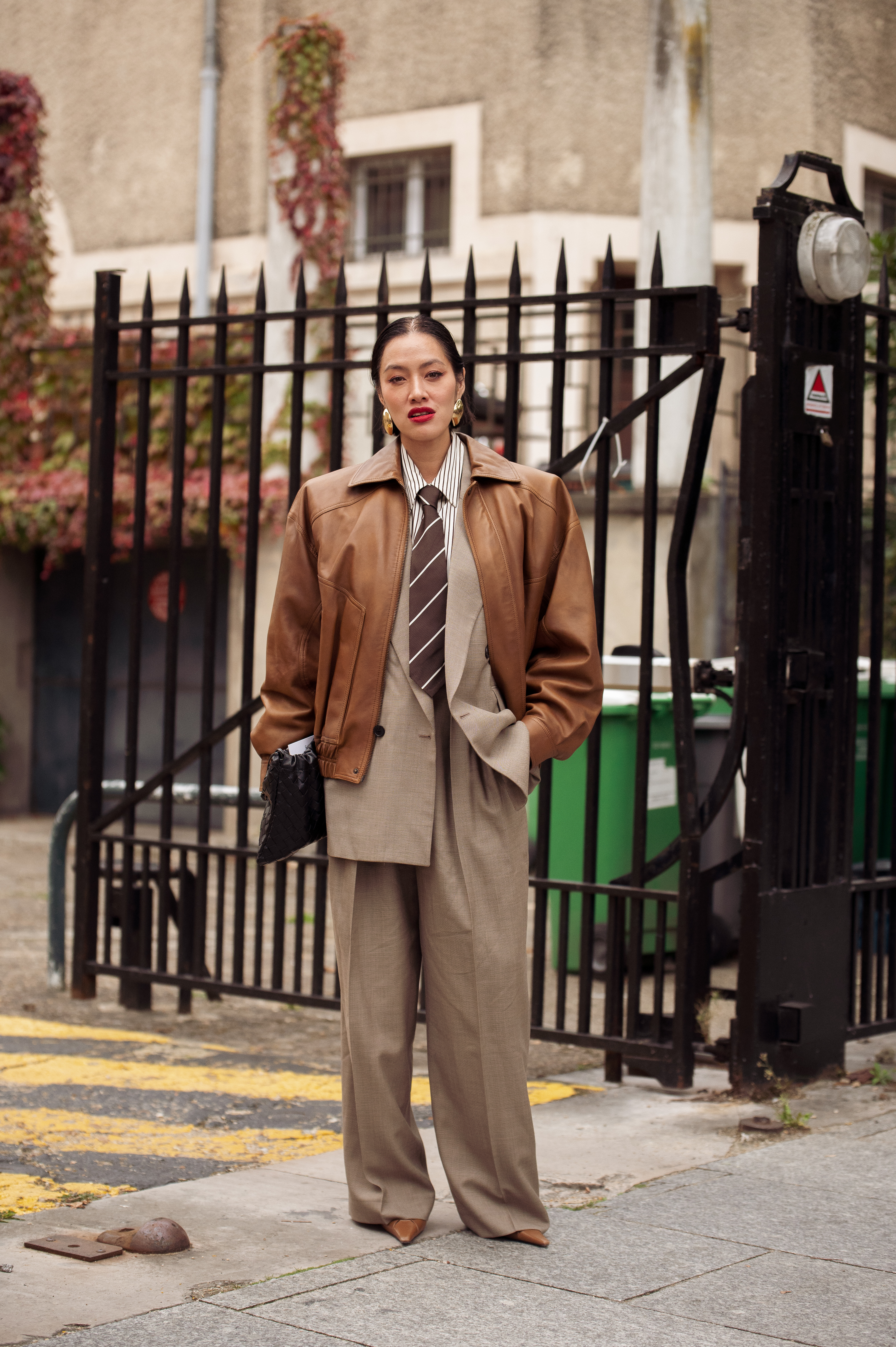 PARIS, FRANCE - OCTOBER 03: Tiffany Hsu wears a taupe oversized suit with a striped shirt and wide tie, layered under a brown leather jacket, styled with pointed tan heels, gold statement earrings, and a black woven clutch outside LOEWE show during the Womenswear Spring Summer 2026 as part of Paris Fashion Week on October 03, 2025 in Paris, France. (Photo by Raimonda Kulikauskiene/Getty Images)