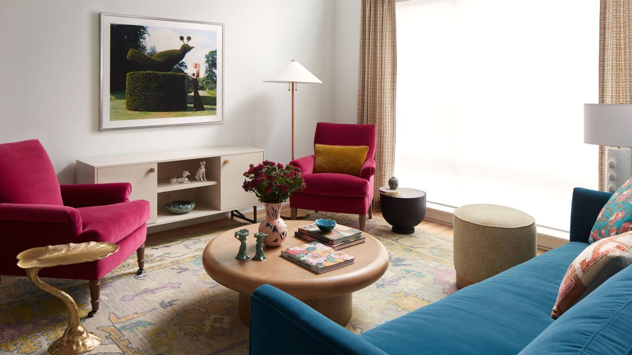 A modern living room with white walls, a beige patterned rug, a round wooden coffee table and a sideboard with a piece of artwork above. Two hot pink armchair and a dark blue sofa in the corner. 