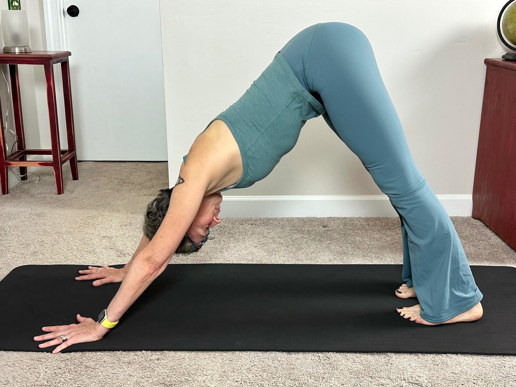 woman wearing blue vest and leggings performing a downward facing dog yoga pose on a black yoga mat in a living room