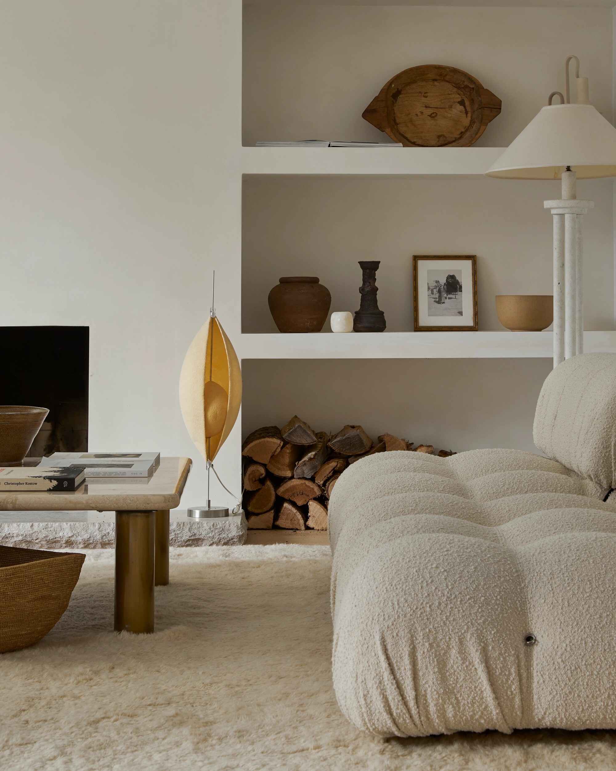 An off-white/neutral but cozy living room. There is open, built-in shelving with wood logs, vases, picture frames, etc. on it. There is a boucle love seat, a fluffy rug, and a marble coffee table.