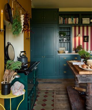 A charming English kitchen featuring deep green cabinetry with brass hardware, complemented by mustard-yellow walls. A dark green AGA cooker sits against a backdrop of vintage-inspired tiles, with hanging herbs and utensils adding rustic appeal.