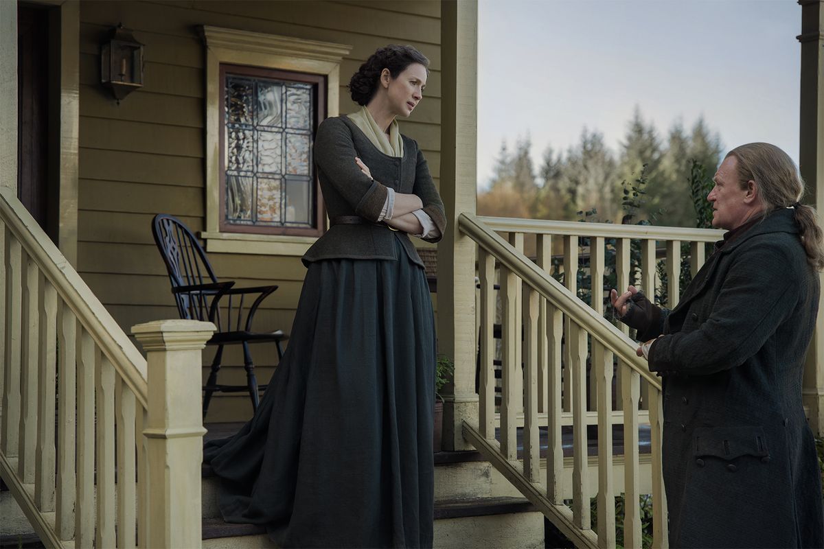 Claire (Caitríona Balfe) stands on the steps outside her home in Fraser&#039;s Ridge, her arms folded and a look of concern on her face, as she meets Tom Christie (Mark Lewis Jones), who stands at the foot of the stairs