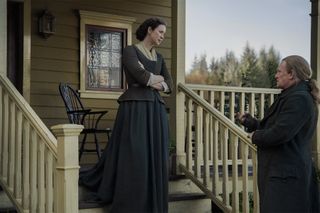 Claire (Caitríona Balfe) stands on the steps outside her home in Fraser's Ridge, her arms folded and a look of concern on her face, as she meets Tom Christie (Mark Lewis Jones), who stands at the foot of the stairs
