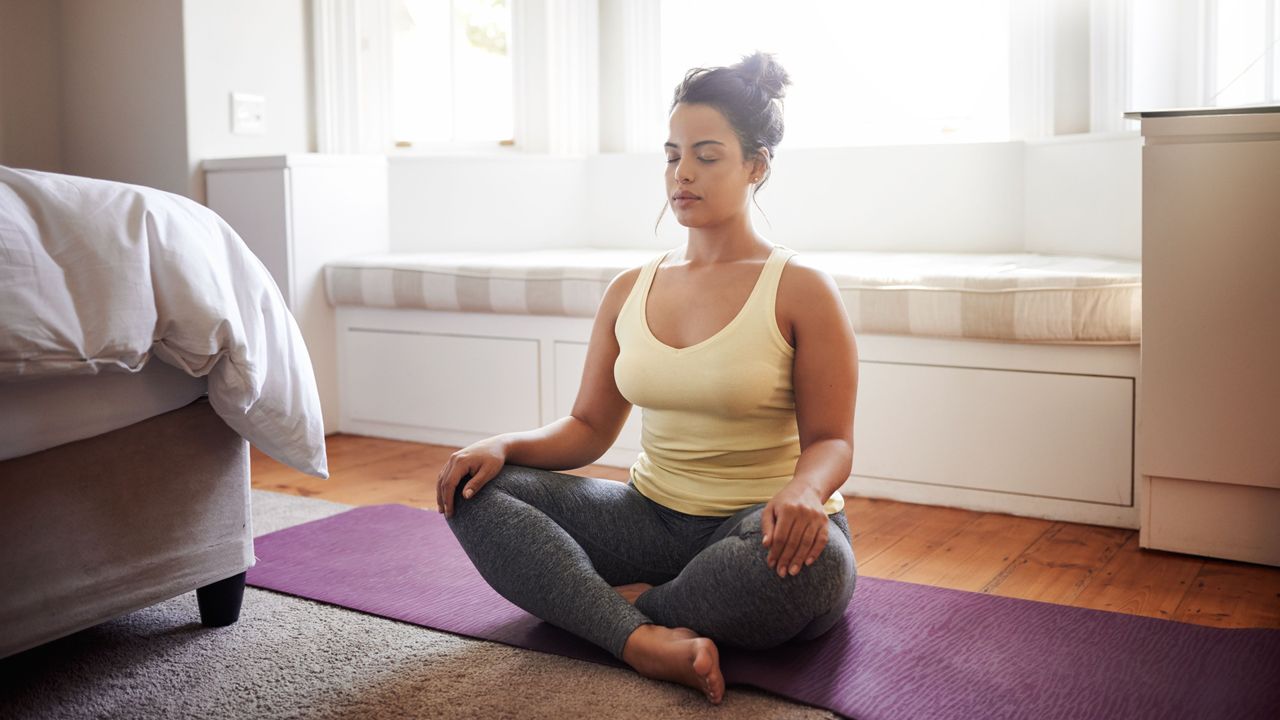 A woman sits cross legged on a yoga mat in her bedroom with her eyes closed