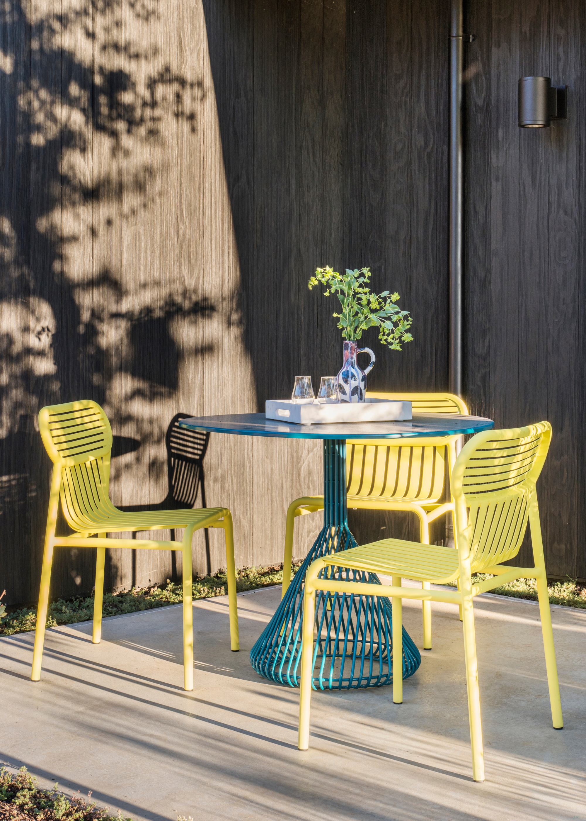 a small outdoor dining area with bold yellow colored seats and a teal table against a backdrop of dark wood clad walls
