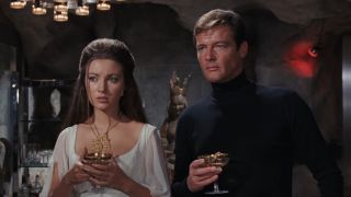 Jane Seymour and Roger Moore stand holding drinks in a lair in Live and Let Die.