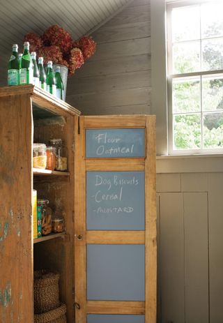 a wooden pantry/larder with doors painted in chalk paint, with food items written on them and food inside the cupboard, and a window to the right