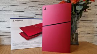 The PS5 Slim with volcanic red console covers on, infront of the console cover box while sitting on wooden table next to a vase of flowers with a white brick background