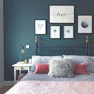 Navy blue wall behind bed with grey patterned bedding and pink cushions with metal frame and prints on the wall