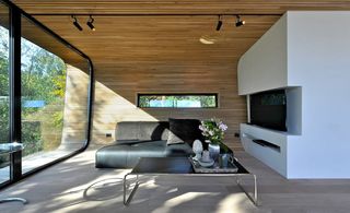 Interior view of the living area at Trekronekabin featuring a wood slat covered ceiling and curved wall, white walls, wood flooring, floor-to-ceiling windows, a tv, a black sofa, a coffee table with a tray of items on top and black bar spotlights