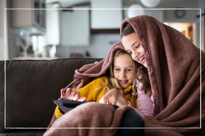 Two children on sofa looking at ipad while snuggled under a blanket