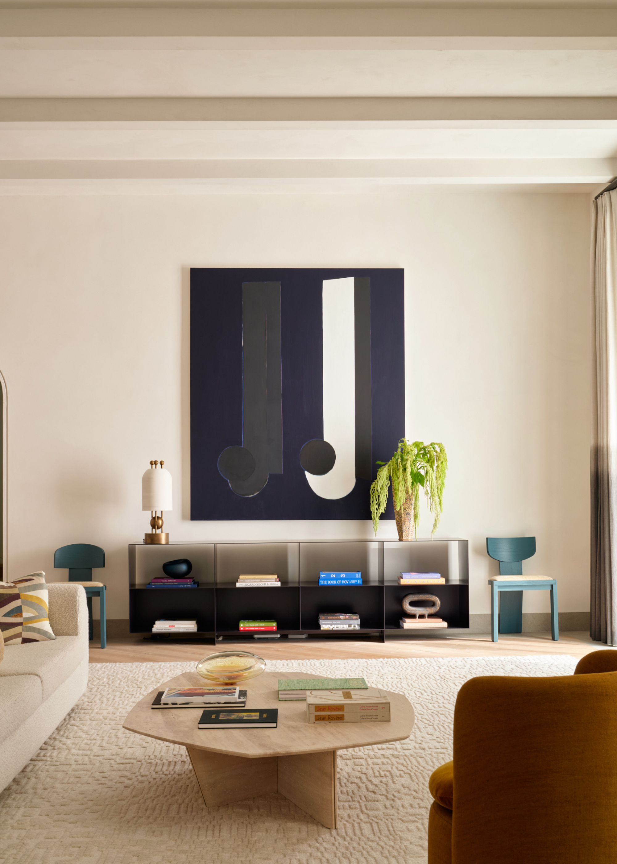 A living room with white walls, a framed black, blue and white abstract painting, above a storage sideboard with colorful books and ceramics, with a vase of green amaranthus, and a gold table lamp, flanked by a pair of blue chairs, in front of a textured off-white rug with a light wood coffee table and an ochre armchair