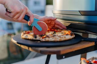 The Gozney x Hedley & Bennett pizza cutter rolling over a fresh pizza beside an outdoor oven