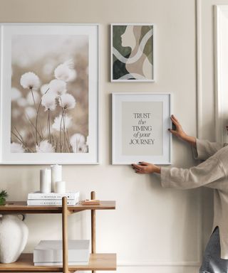 A beige wall with three wall art prints, with one being hung up by a person with a gray shirt