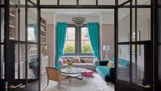 Living room seen through black-framed glass doors, grey and teal velvet sofas, teal curtains, chandelier.