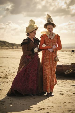 Two women stood on a beach in period dresses and hats