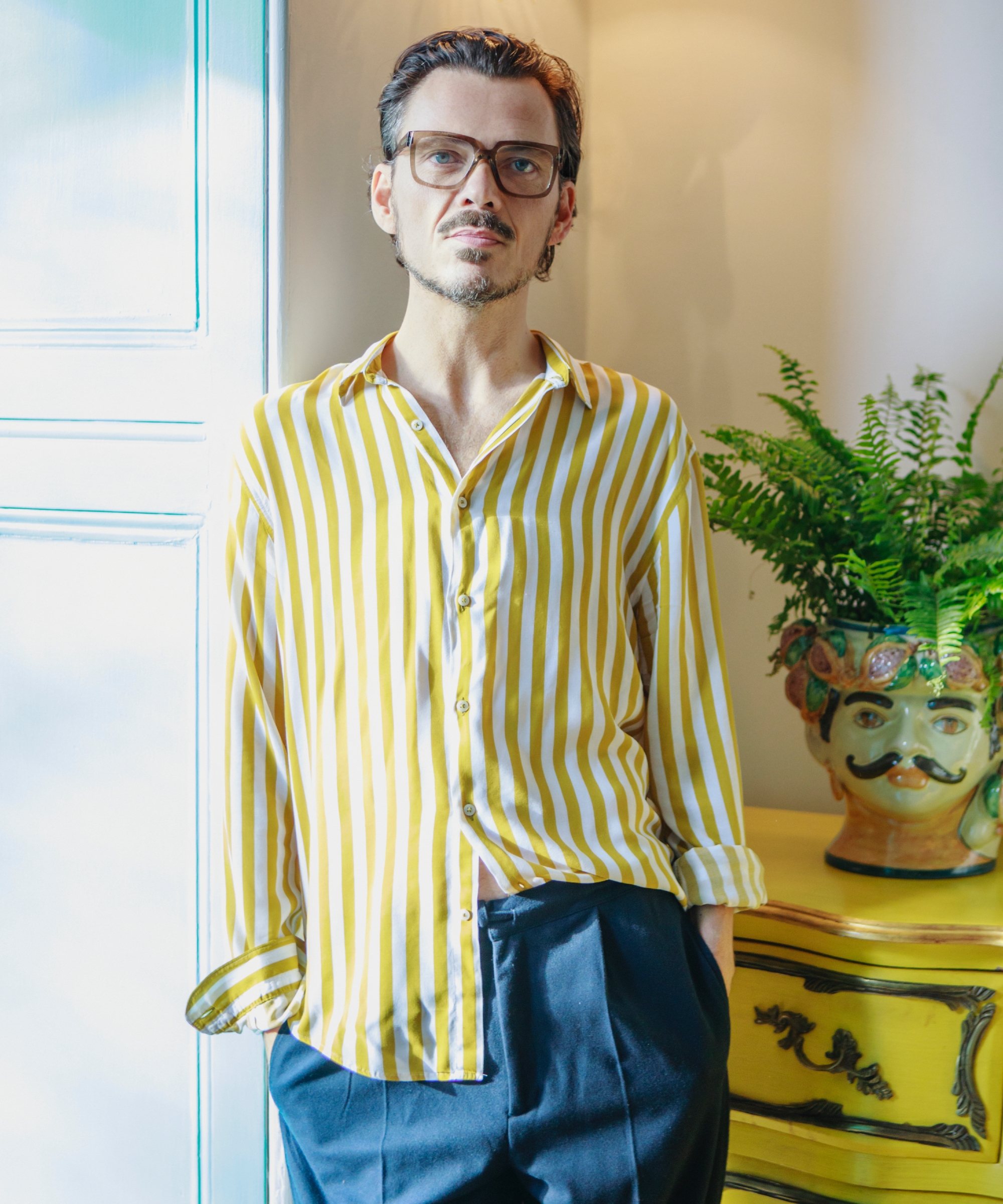 image of Matthew Williamson, global interior designer standing by a window wearing a yellow and white stripe shirt, blue trousers, standing in front of yellow console table