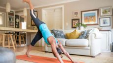 woman wearing a turquoise vest doing a downward facing dog with one leg raised on an orange exercise mat in a living room setting. there's a breakfast bar and a cream sofa behind her.