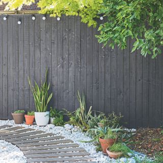 Black painted fence above a walkway consisting of white stones and wooden planks, with potted plants next to it