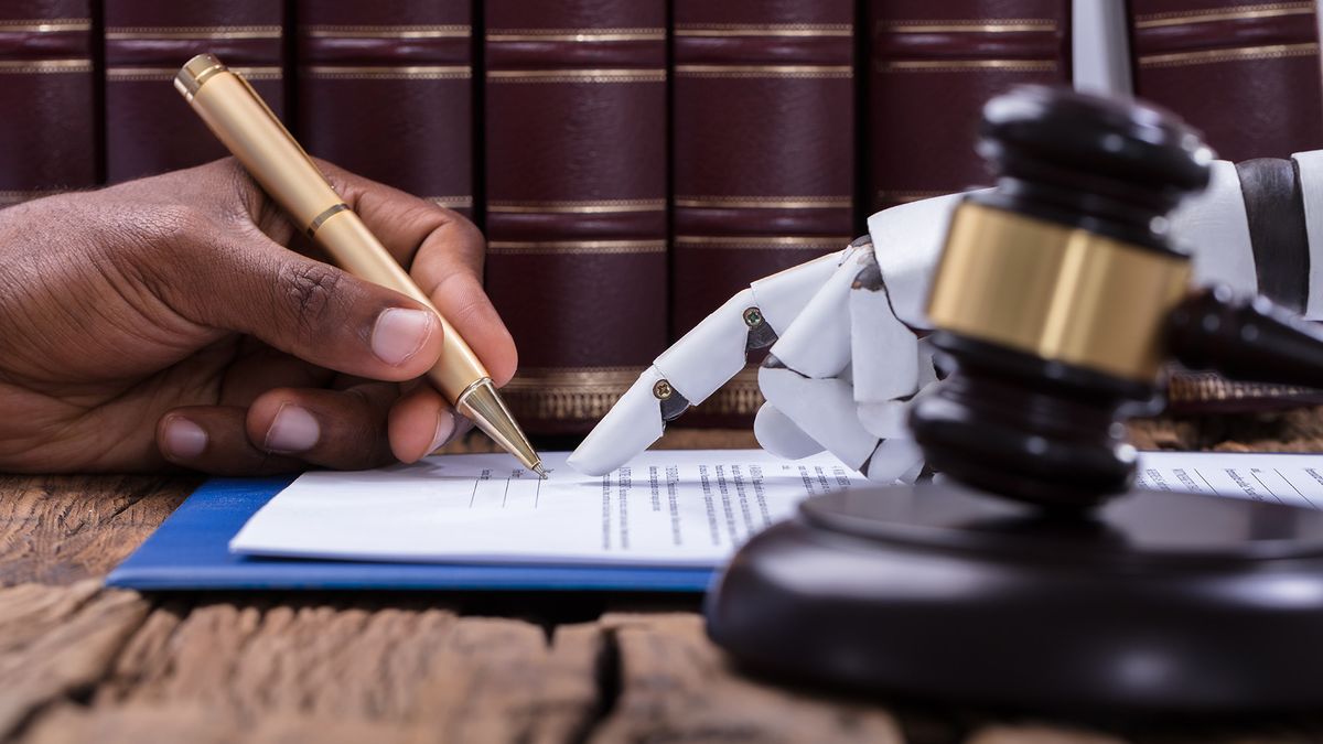 A photo of a person&amp;#039;s hand signing a document with a robot&amp;#039;s hand pointing at where to sign. There is a gavel in the foreground on the robot&amp;#039;s side of the table.