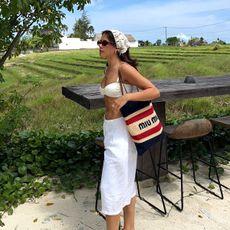 Influencer Tia Dewitt wearing a Miumiu knit tote, linen white pants, a white bikini top, and rope sandals. 