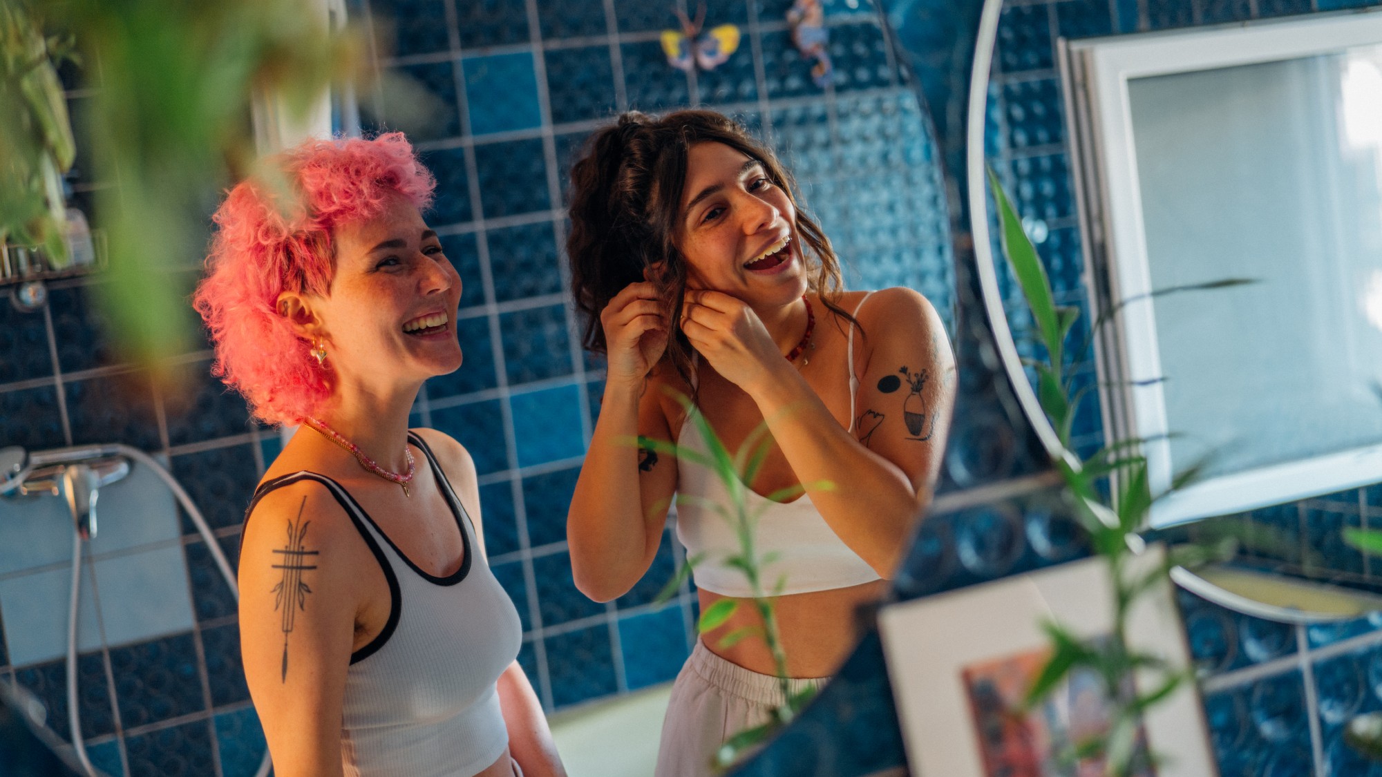 two women getting ready in front of a mirror. both have tattoos. one has pink wavy hair, the other dark hair. they might be dating