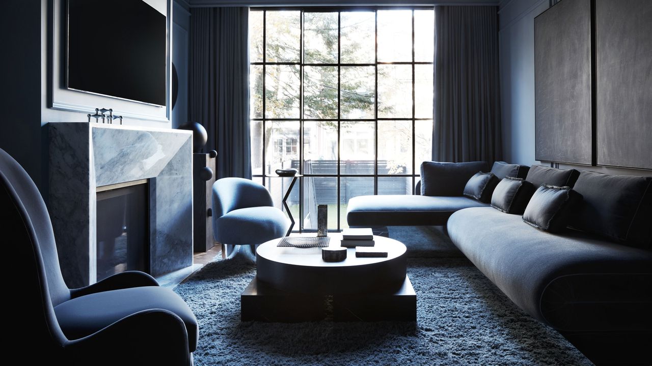 Image of a blue living room with a sofa, two accent chairs, and a circle coffee table. There is a large tv hanging over the fireplace