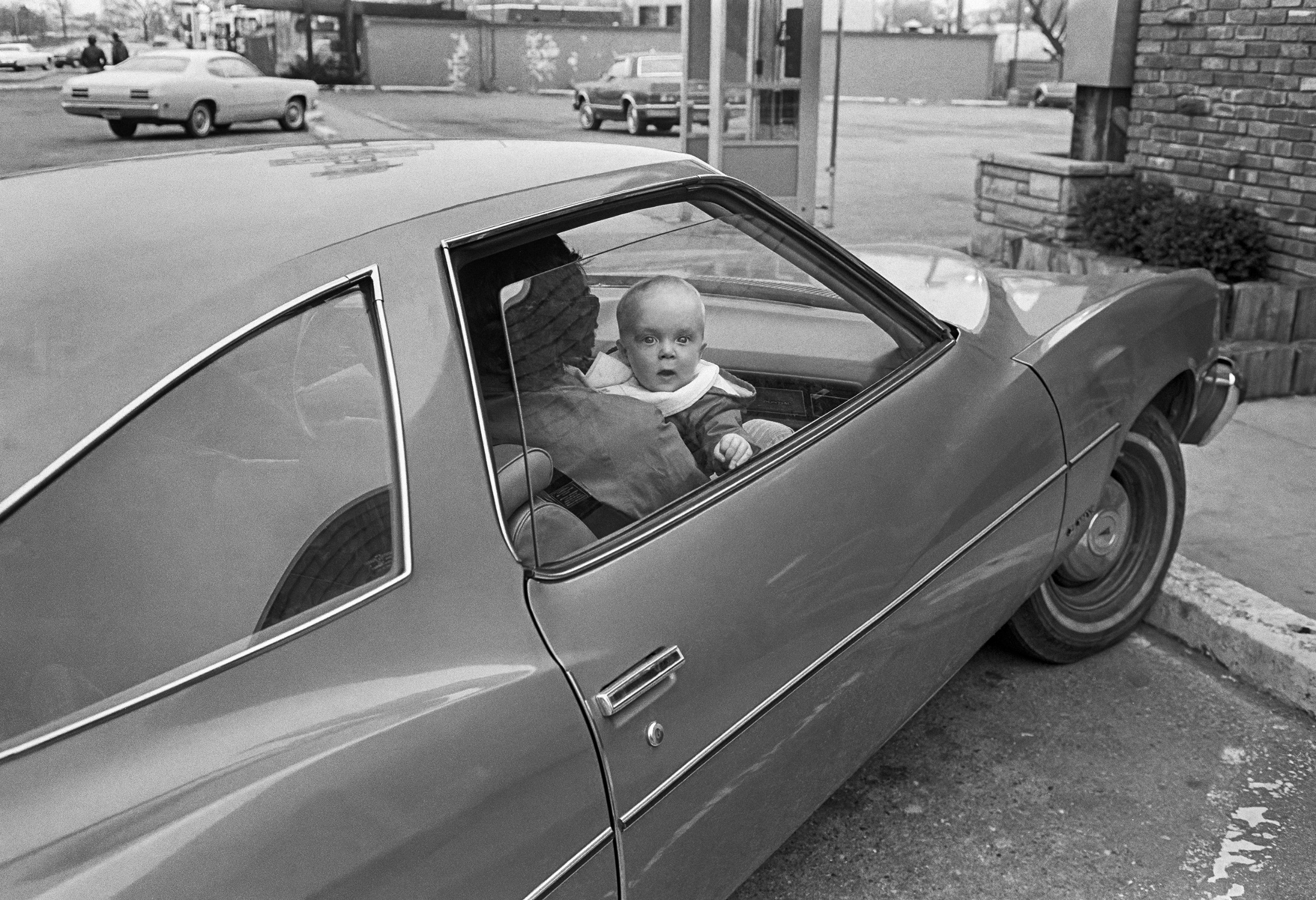 A wide-eyed baby held by an adult peers out of the window of a parked car, with a car park and other vehicles in the background.