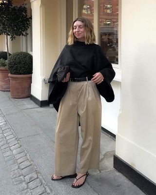 British female fashion influencer Brittany Bathgate poses on a Copenhagen sidewalk weainf a black cape top, black clutch bag, black belt, tan wide-leg cropped pants, and strappy black flat sandals