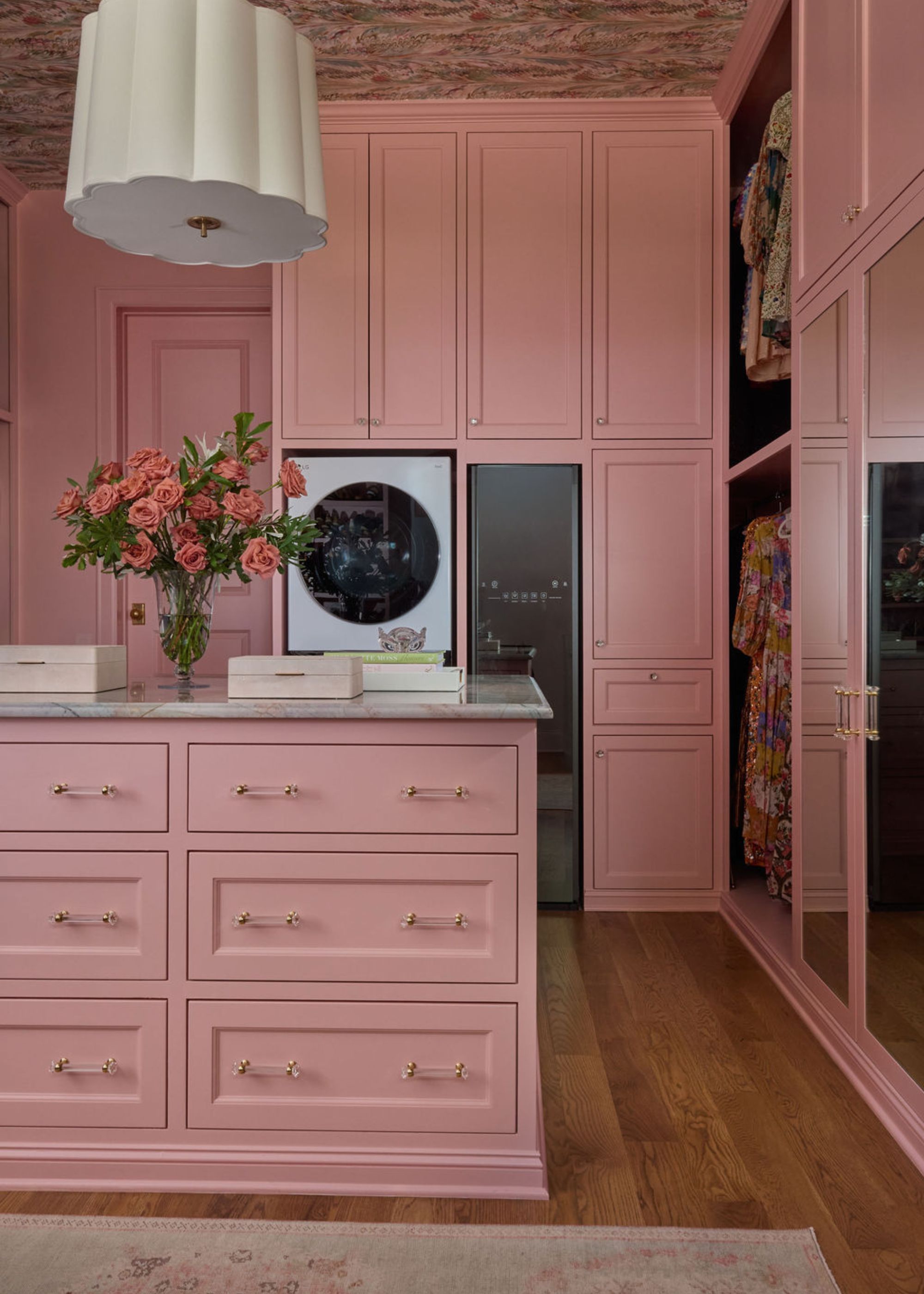 Large dressing room with full-height cabinetry, color-drenched in a muted rose color, and a matching island unit. Patterned wallpaper on the ceiling in pink tones, wooden flooring, and a pendant light