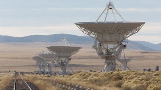 four radio telescopes along a railroad track up in a scrub desert pointin straight all up in tha sky. mountains is up in tha distance