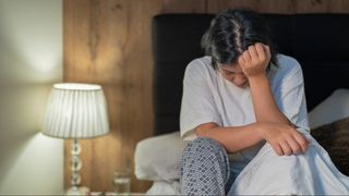 A woman in white top and pyjama bottoms sitting up in bed with her head in her hands.