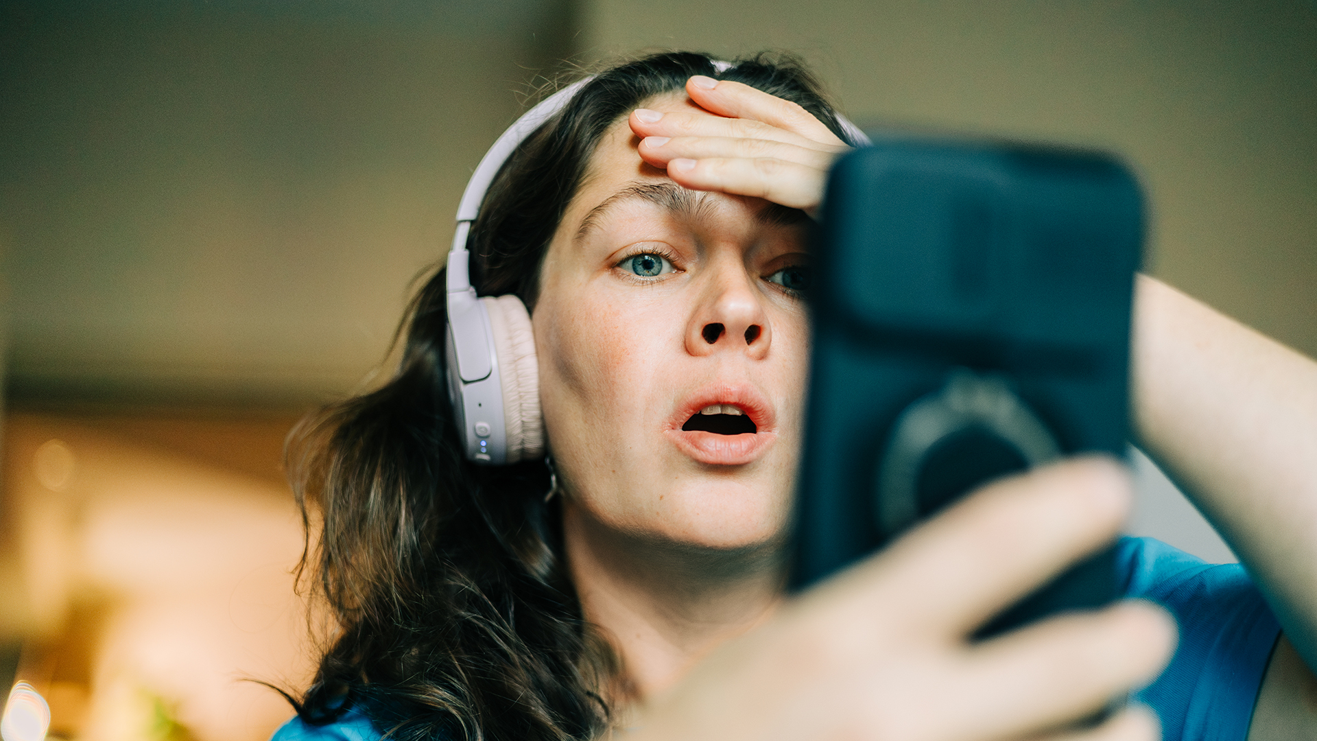 A woman in headphones looks shocked at her phone, holding her hand to her head