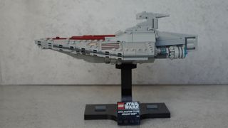 A miniature-sized long grey ship made of bricks with a red stripe running down the middle is placed on a black stand in front of a grey background.