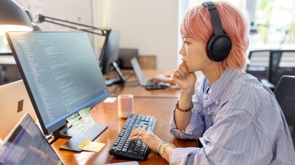Female software developer with pink hair and headphones conducting software testing on a desktop computer in an open plan office space.