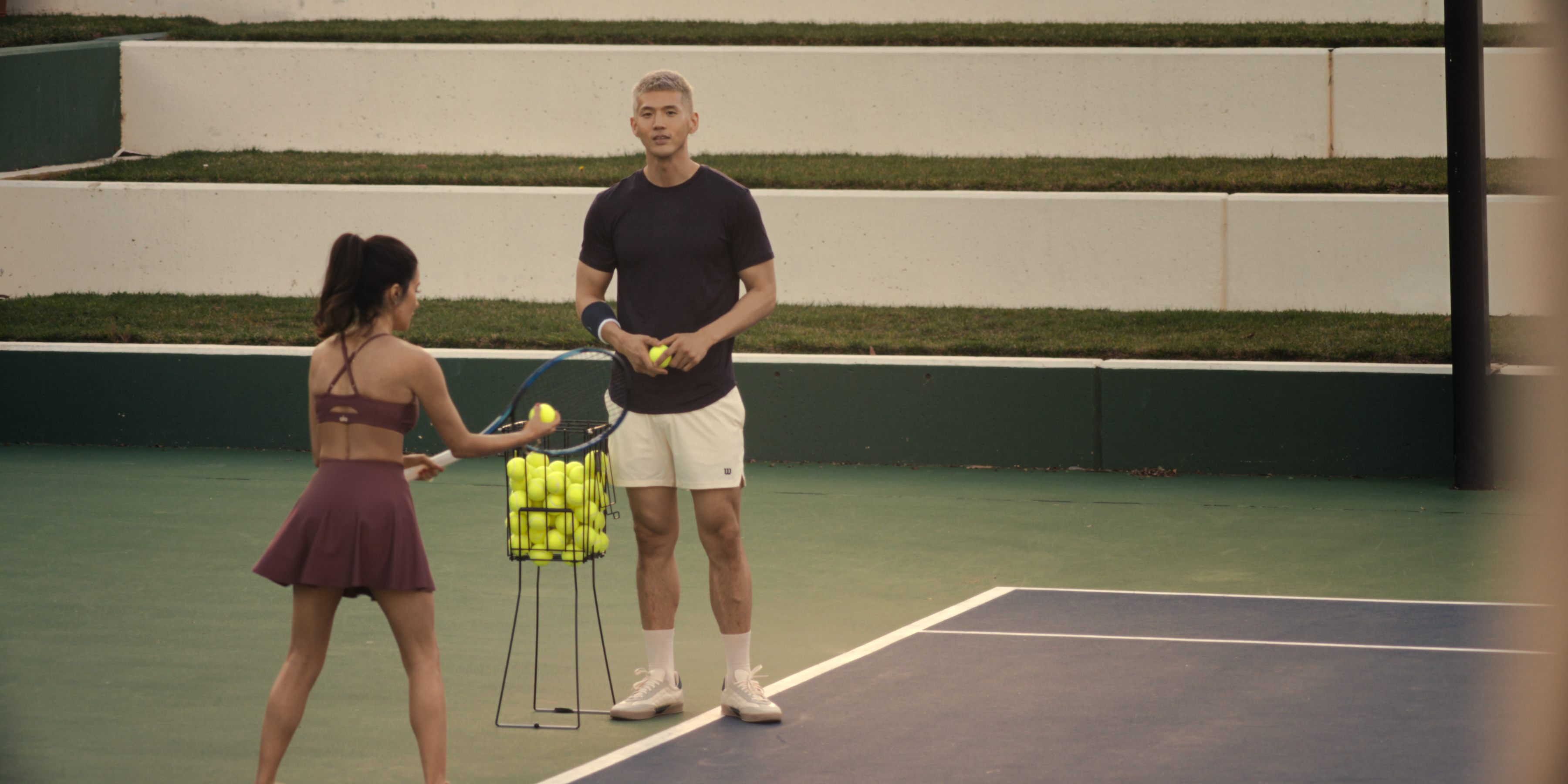 A woman in a tennis outfit (Mikaela Hoover as Ava) prepares to serve as a man holding a ball (Matthew Kim as Woosh) stands nearby on a tennis court, in episode 201 of 'Beef.'