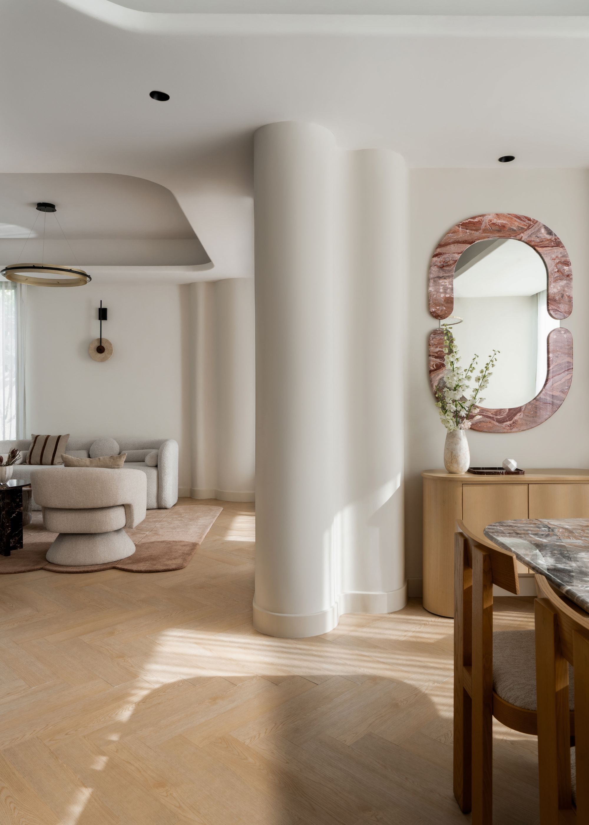 A white open space beside the dining room and living space with curved pillars, a stone framed mirror, a marble dining table, wood chairs, and minimalist living room furniture