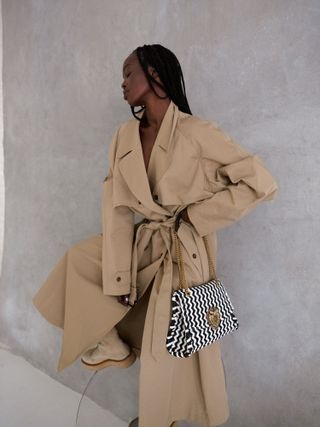 A model wearing a tan Dolce and Gabbana trench coat in front of a gray wall.