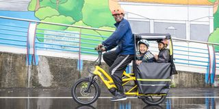 A man rides a yellow Tern GSD cargo bike with two young children sat at the rear covered by a rain tent.